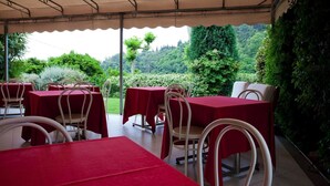 An outdoor dining area with tables covered in red tablecloths and white chairs, set against a backdrop of lush greenery and a view of a forested hill. - Hotel Ristorante Grappolo D'Oro (Pietrasanta)