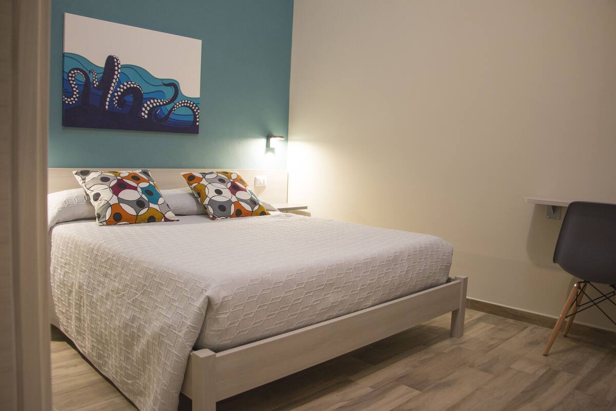 A neatly made bed with a white quilt and colorful pillows, a wall-mounted light, and a chair with a dark seat and wooden legs.