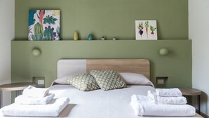 Una cama bien tendida con sábanas blancas y dos almohadas estampadas, una pared verde con obras de arte y una mesita de noche con toallas dobladas.