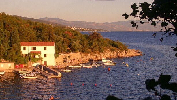 Un edificio costero con techo rojo, barcos amarrados y vista al mar y montañas lejanas.