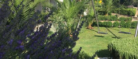 A garden with lavender and palm trees, a swing set, and a building with a balcony.