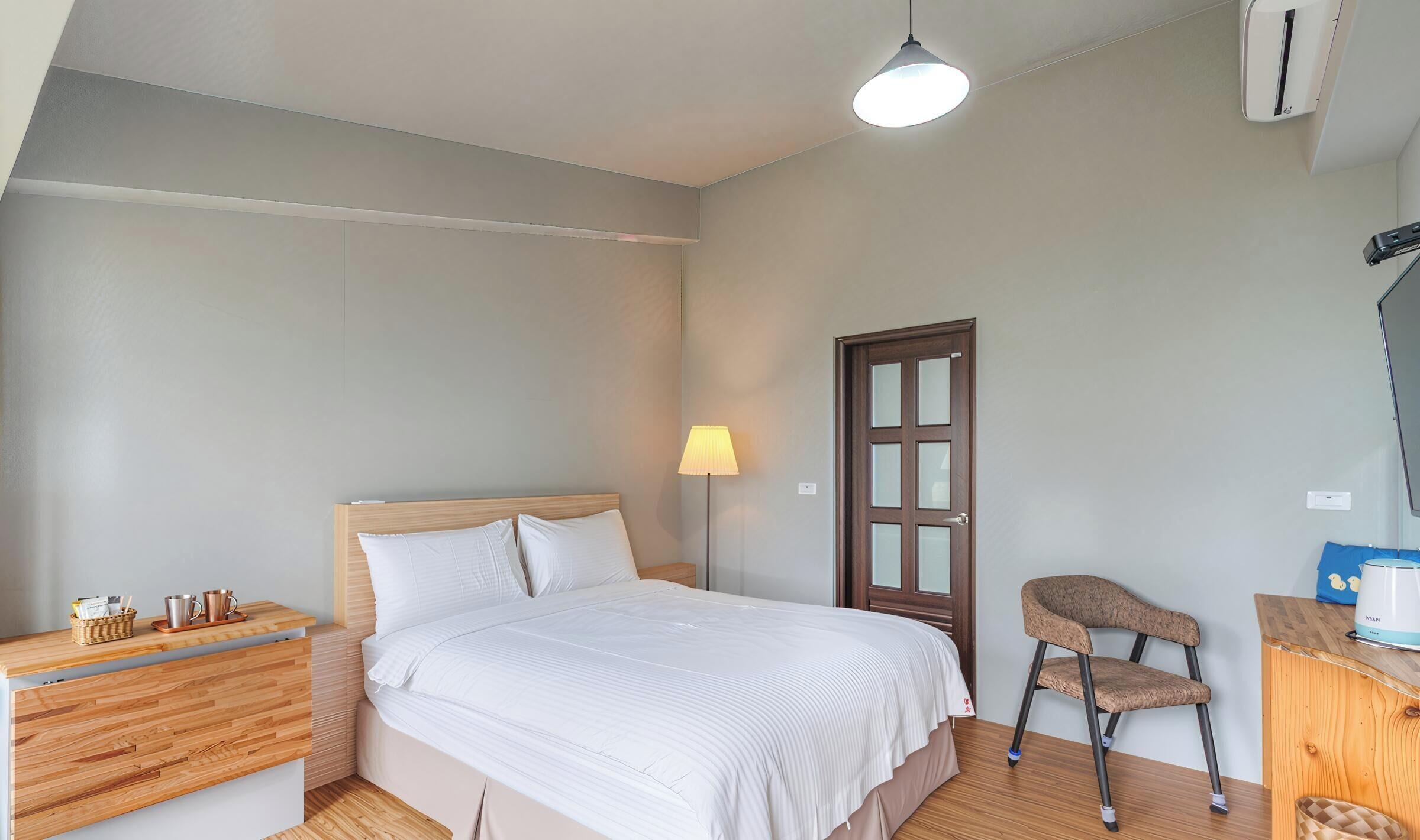 A bedroom with a bed, bedside table, lamp, chair, and a desk with a kettle.
