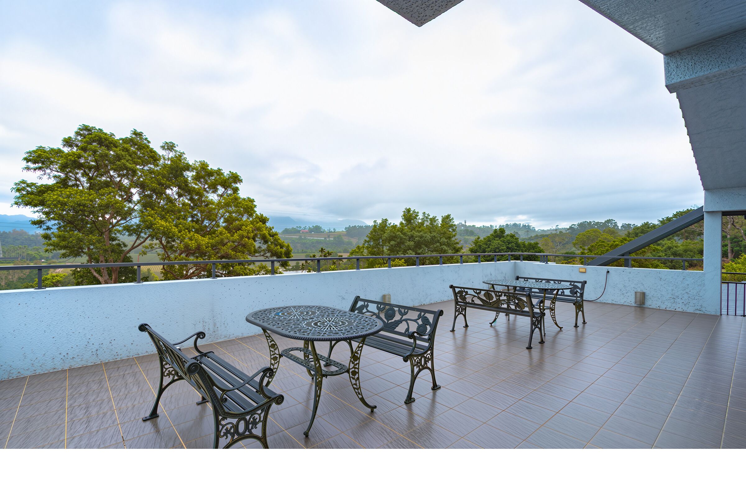 Ein Balkon mit rundem Tisch, vier Stühlen, Geländer und Blick auf Bäume und Hügel.