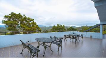 Ein Balkon mit rundem Tisch, vier Stühlen, Geländer und Blick auf Bäume und Hügel.