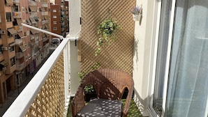 Un balcón con una silla de mimbre, una planta en maceta y vistas de edificios cercanos.