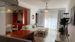 Una sala de estar moderna con un sofá, una mesa de centro y un comedor con una mesa y sillas rojas. Hay una planta en maceta sobre una mesita auxiliar y un ventanal con cortinas.