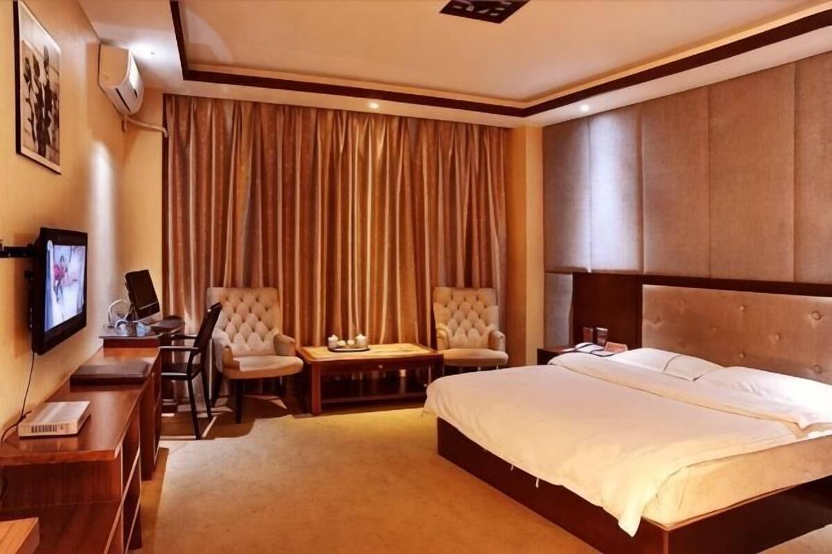 A hotel room with a large bed, two armchairs, a desk with a computer, and a TV.