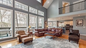 A spacious living room with large windows, a cozy sectional sofa, and a dining area with a kitchen in the background. - Luxury Lakefront w/ Hot Tub & Rec Room (Oakland)