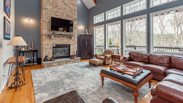A spacious living room with a stone fireplace, leather furniture, and large windows offering a view of trees. - Luxury Lakefront w/ Hot Tub & Rec Room (Oakland)