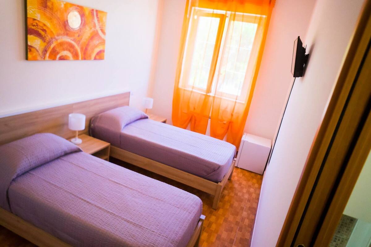 Ein Hotelzimmer mit zwei Einzelbetten, einem Nachttisch mit Lampe, einem Fenster mit durchsichtigen Vorhängen und einem an der Wand befestigten Fernseher.