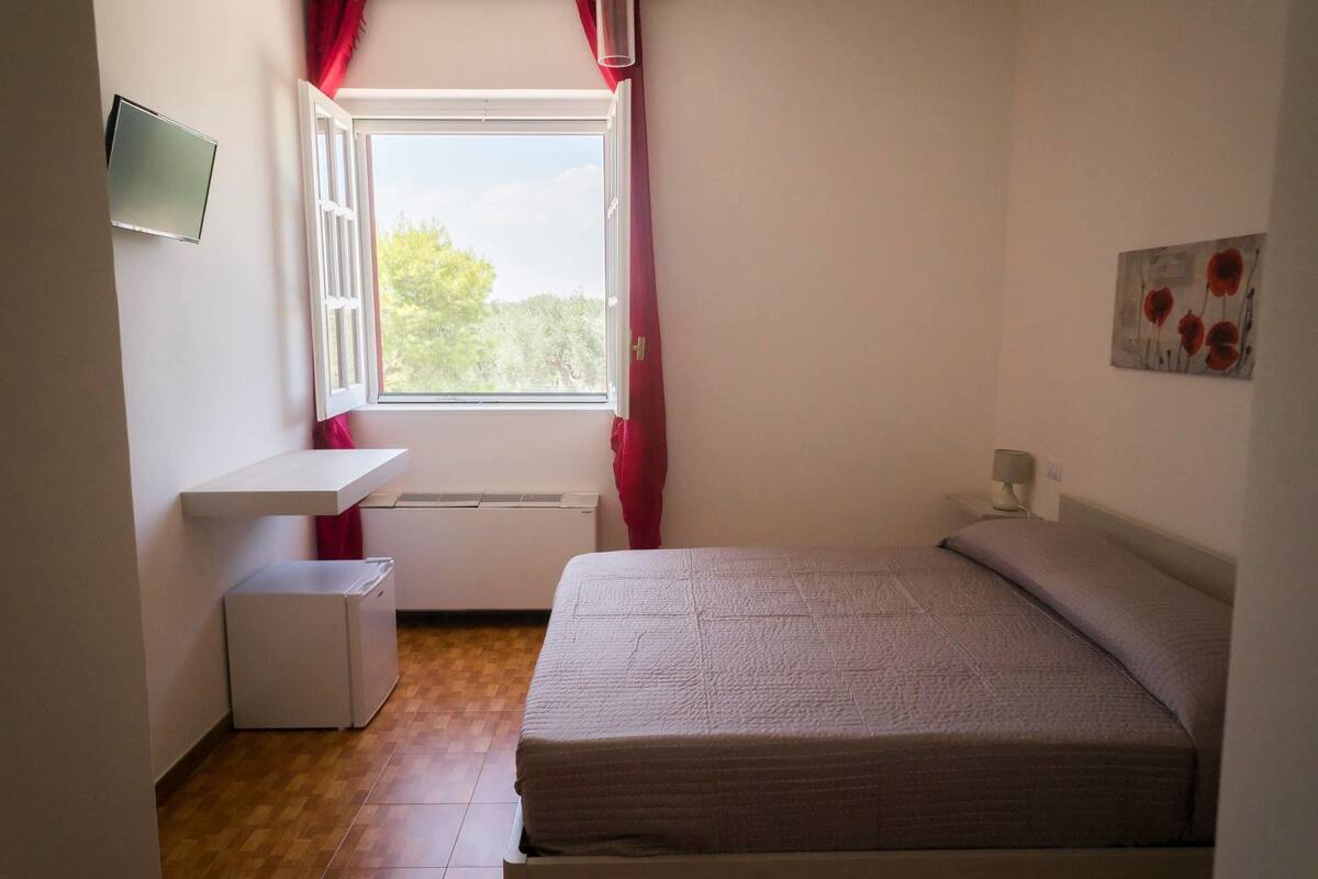Ein Schlafzimmer mit Bett, Nachttisch, Fenster mit Vorhängen, Heizkörper und einem am Wand montierten Fernseher.