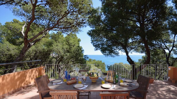 A round wooden table set for dining with chairs, a basket of flowers, and a view of the sea. - Holiday rental villa with swimming pool in Begur, Sa Tuna (Begur)