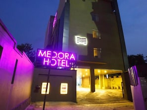 A hotel entrance with a neon sign, a modern building facade, and a paved driveway leading to the entrance. - Medora Hotel (Kozhikode)