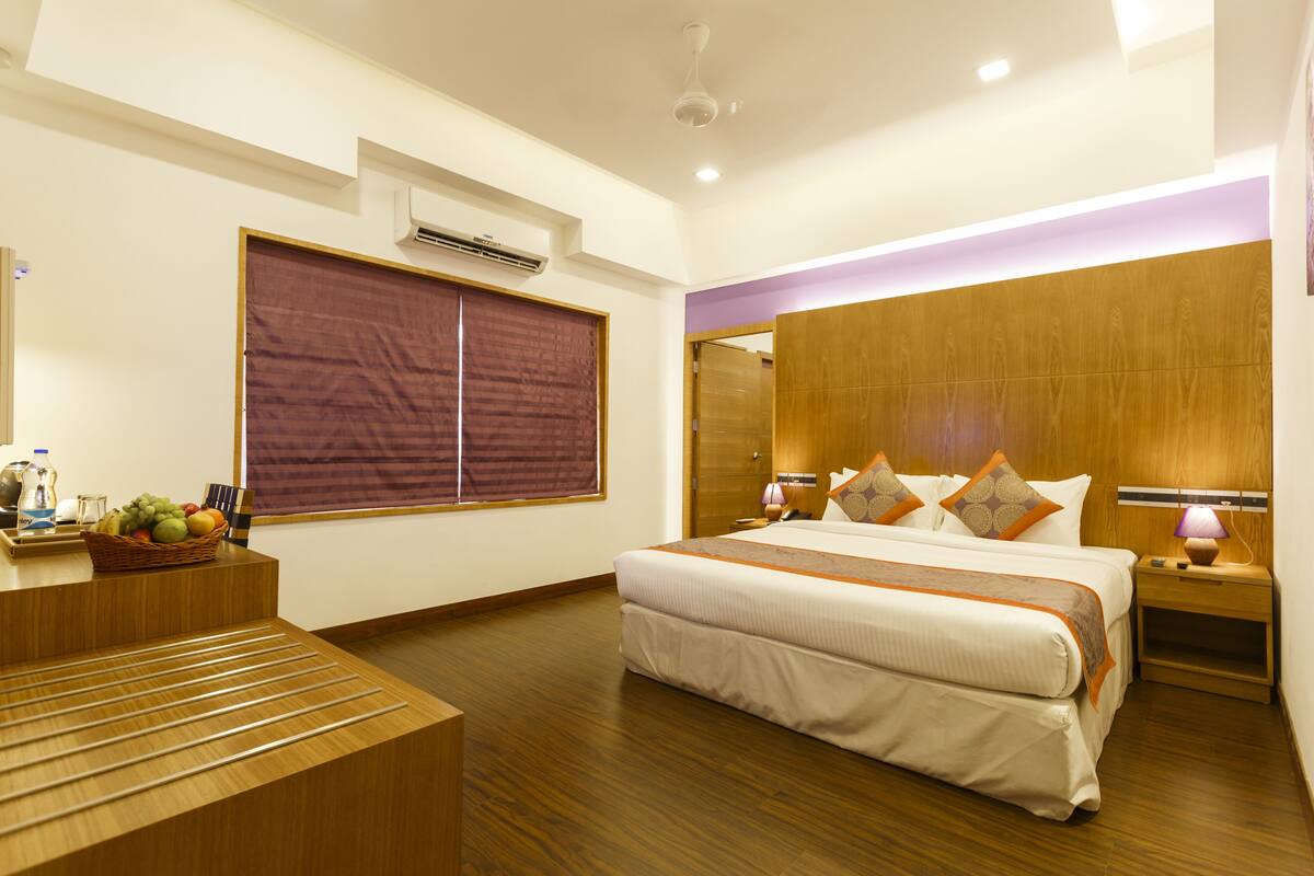 A modern hotel room with a large bed, wooden flooring, a wooden headboard, a bedside table with a lamp, and a window with curtains.