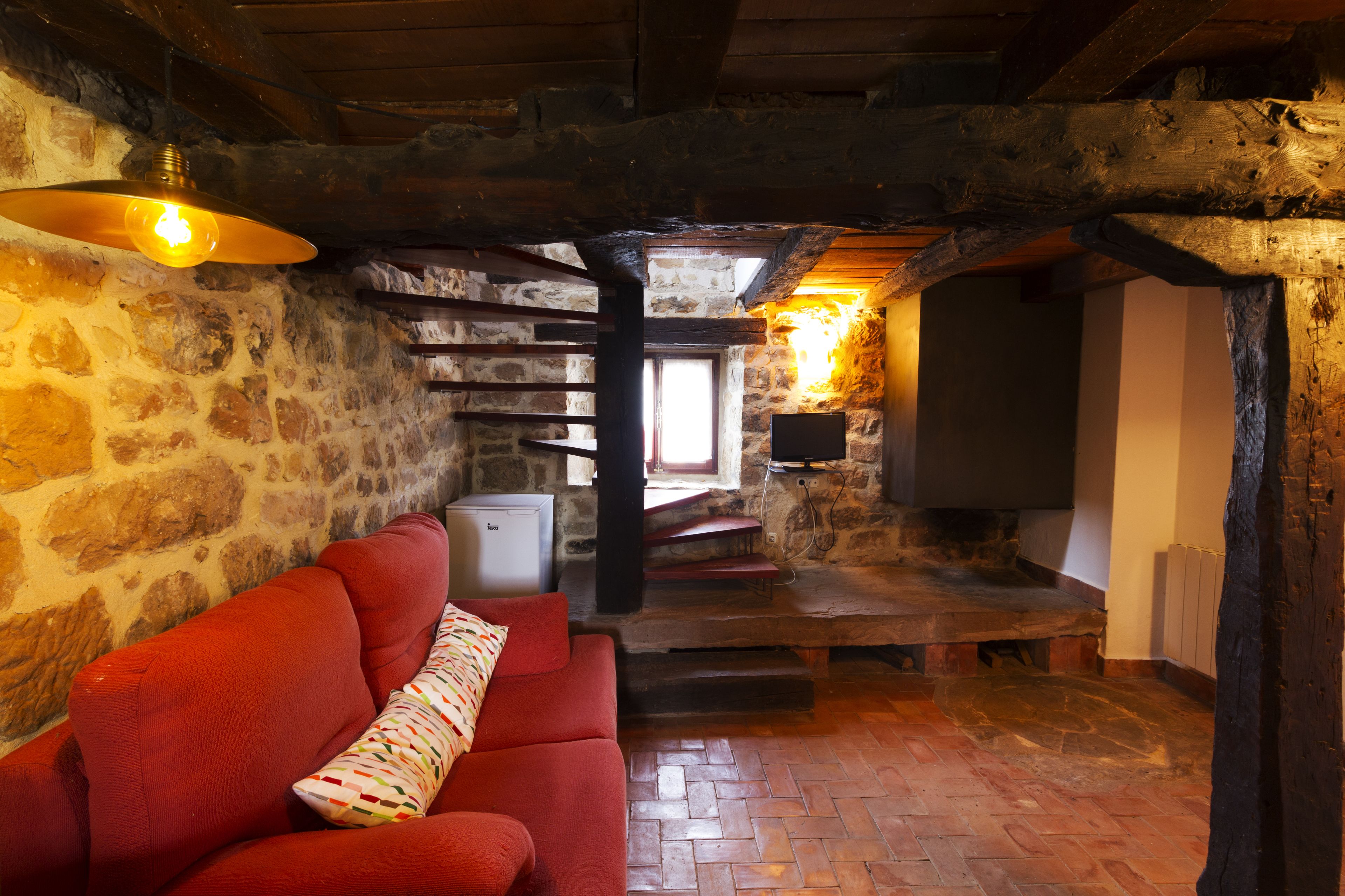 A cozy room with a red sofa, stone walls, wooden beams, a fireplace, and a television.