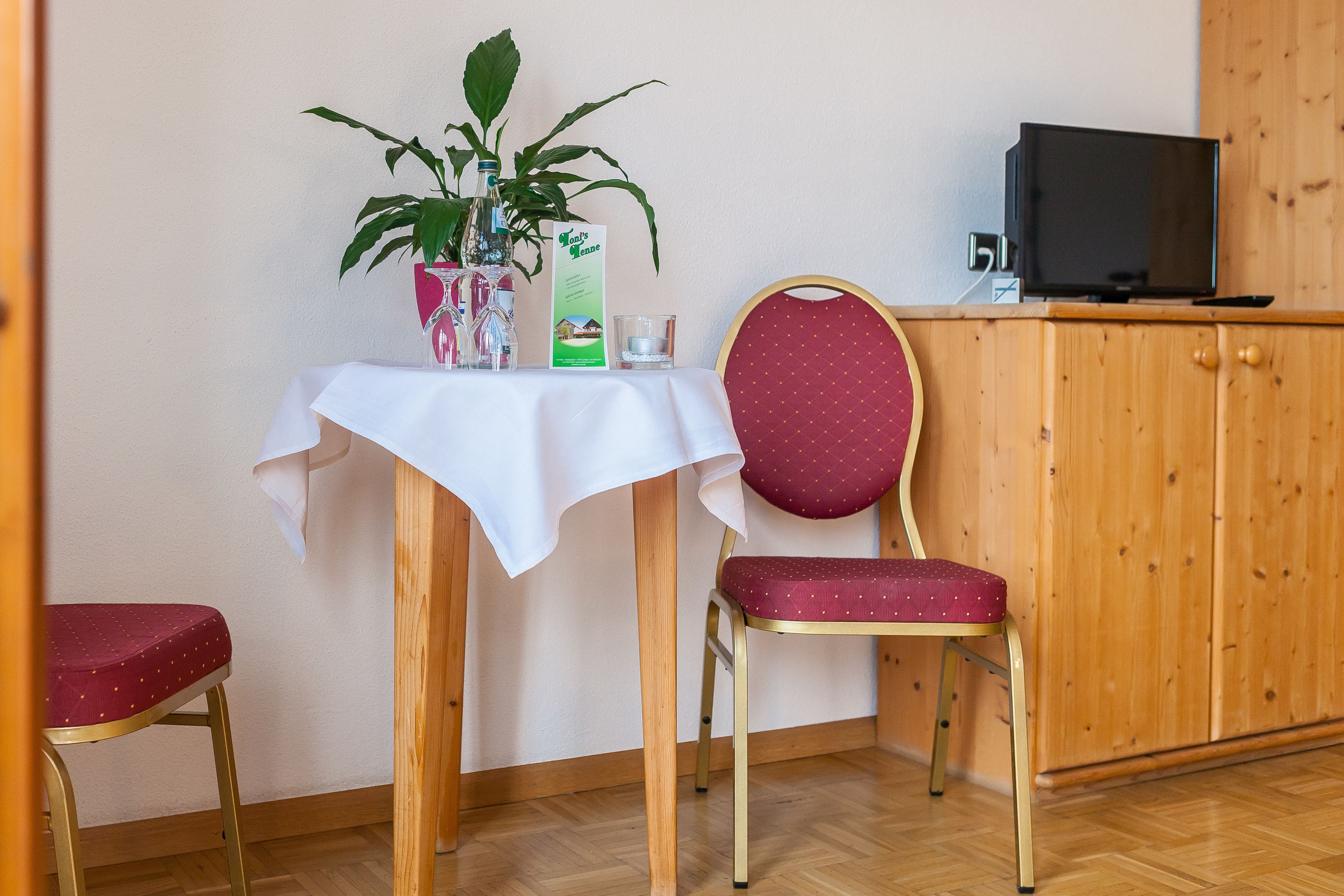 A small room with a wooden cabinet, a television on top, a table with a white tablecloth, a chair with a red cushion, and a potted plant.
