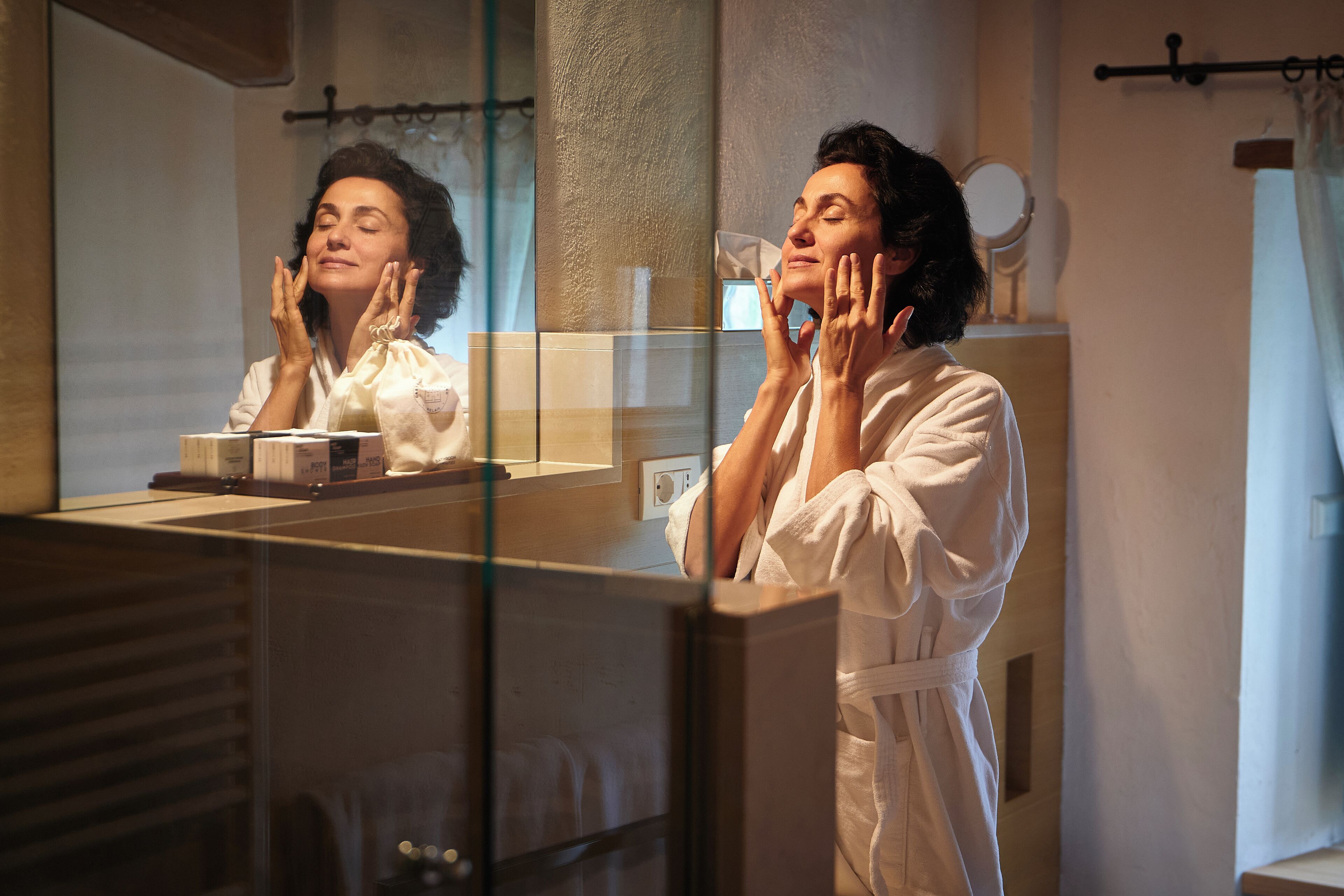 A person in a white robe standing in front of a bathroom mirror, with a towel rack and a small round mirror on the wall.
