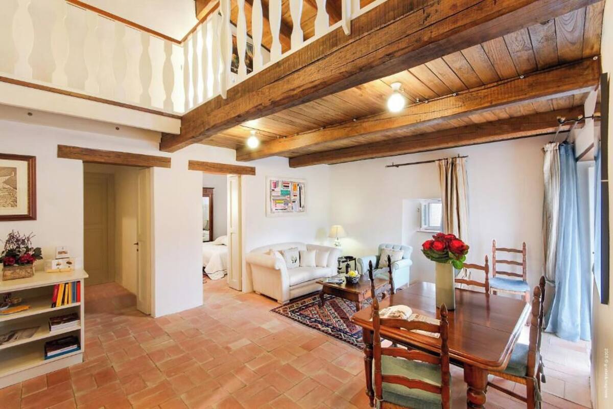 A spacious living area with a high ceiling, wooden beams, a dining table with chairs, a sofa, a bookshelf, and a decorative vase with flowers.
