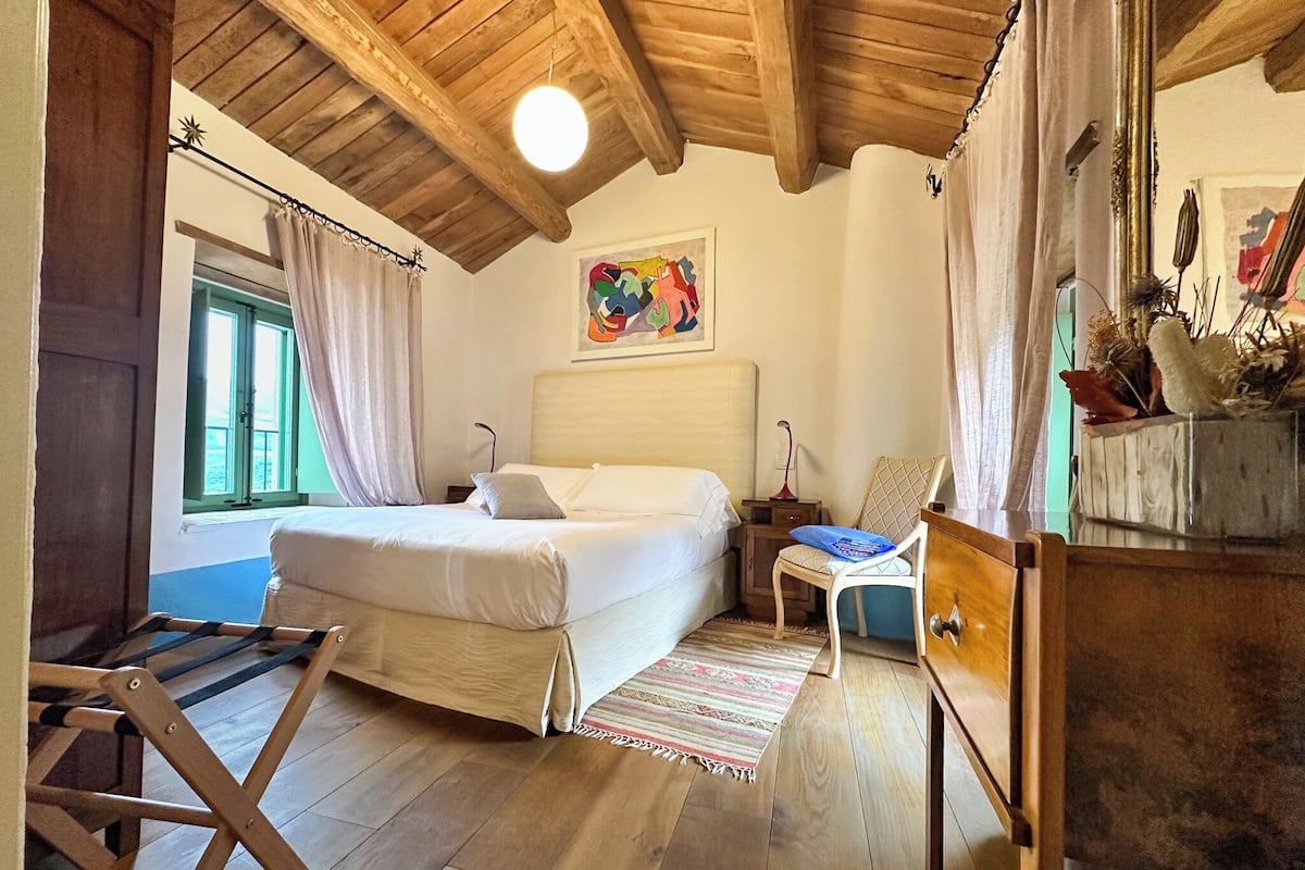 A bedroom with a wooden ceiling, a bed, a nightstand, a chair, and a window with curtains.