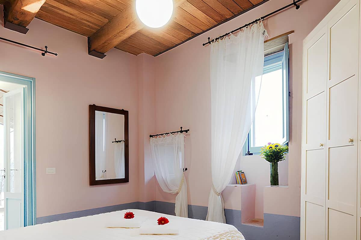 A bedroom with a wooden ceiling, a bed with a white duvet, a mirror, a window with white curtains, and a small vase with flowers.