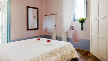 A bedroom with a wooden ceiling, a bed with a white duvet, a mirror, a window with white curtains, and a small vase with flowers.