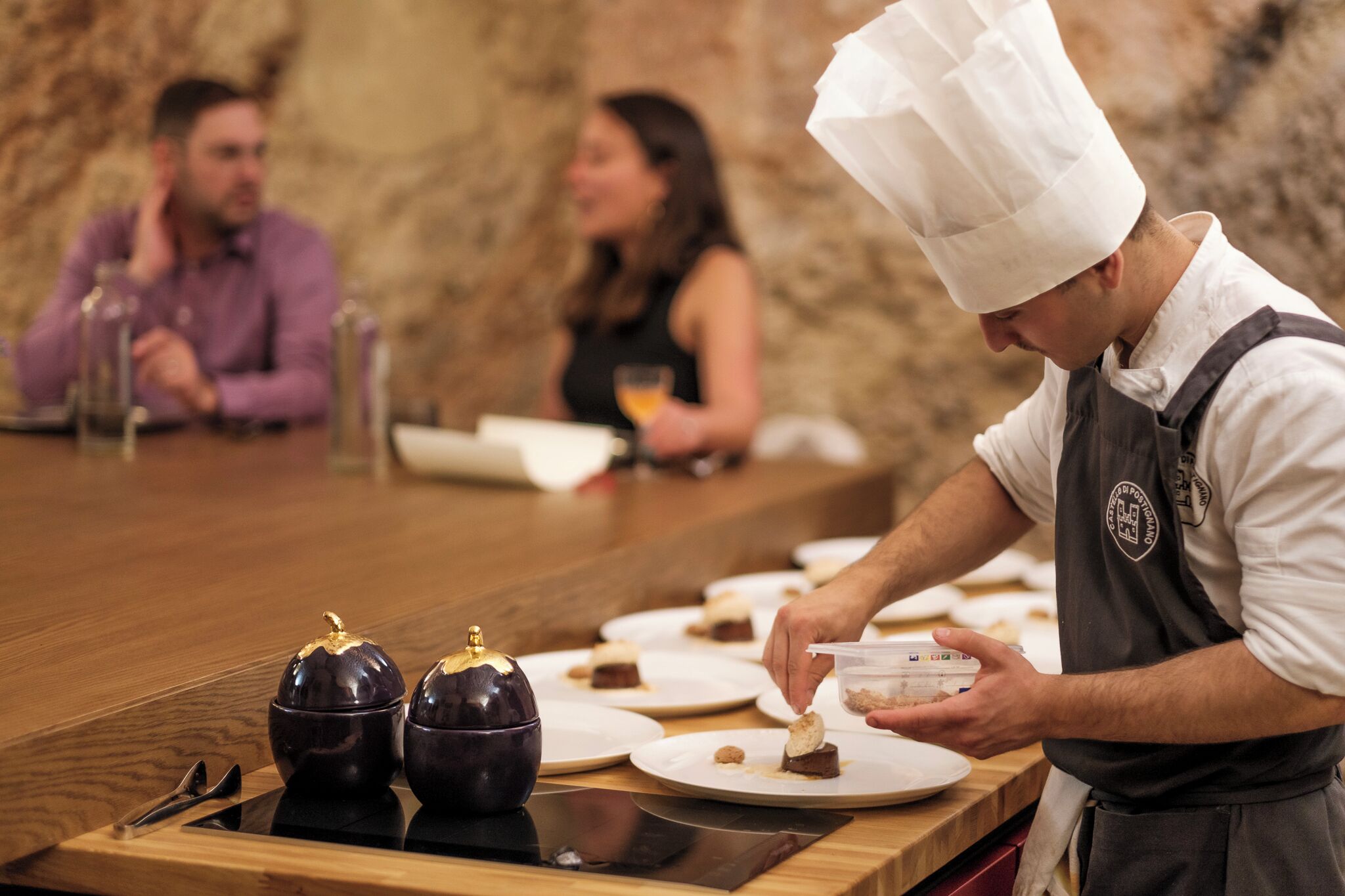 A chef serving food at a counter with a stone wall background.