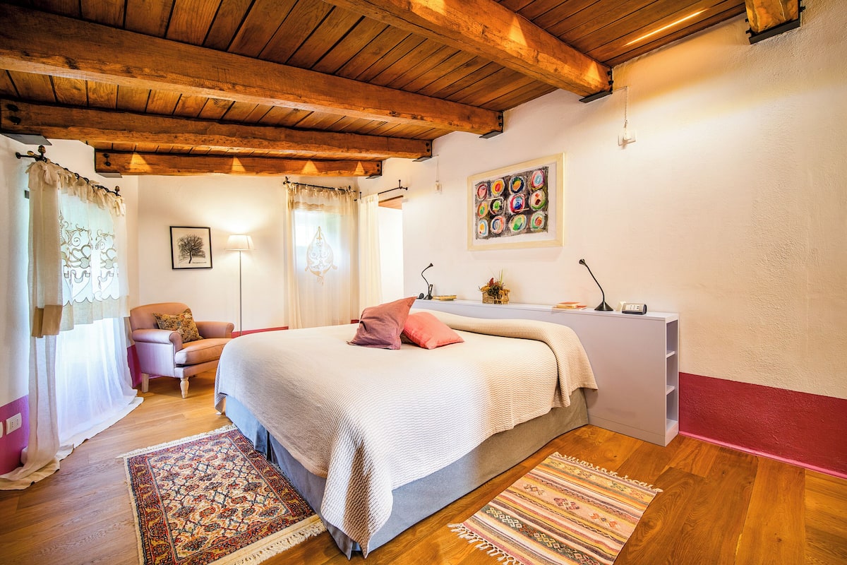 A bedroom with a wooden ceiling, a bed with a white and pink blanket, a bedside table with a lamp, and a wall-mounted picture.