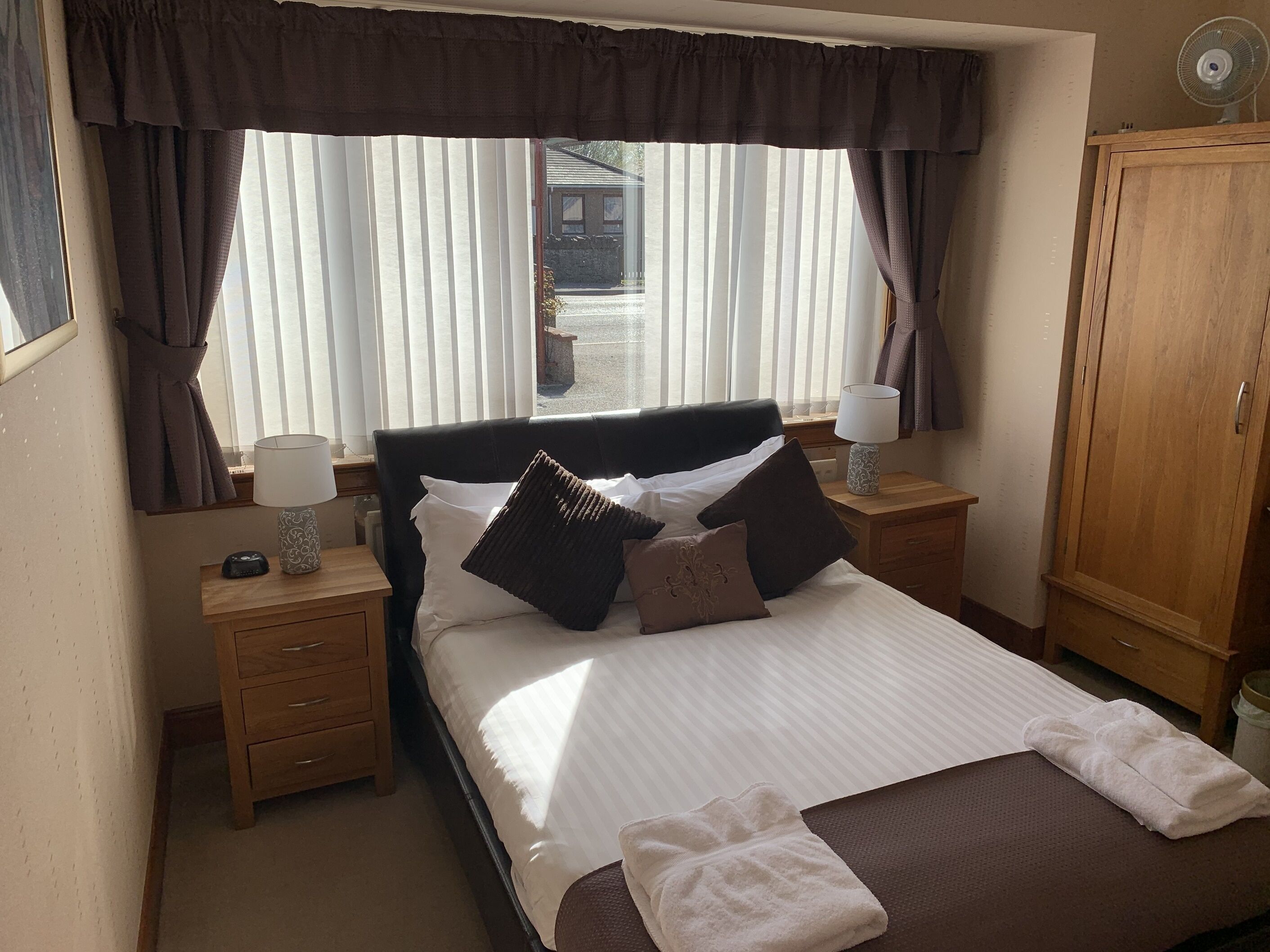 A neatly made bed with pillows and a blanket, a bedside table with a lamp, a wooden nightstand, a wooden wardrobe, and a window with curtains.