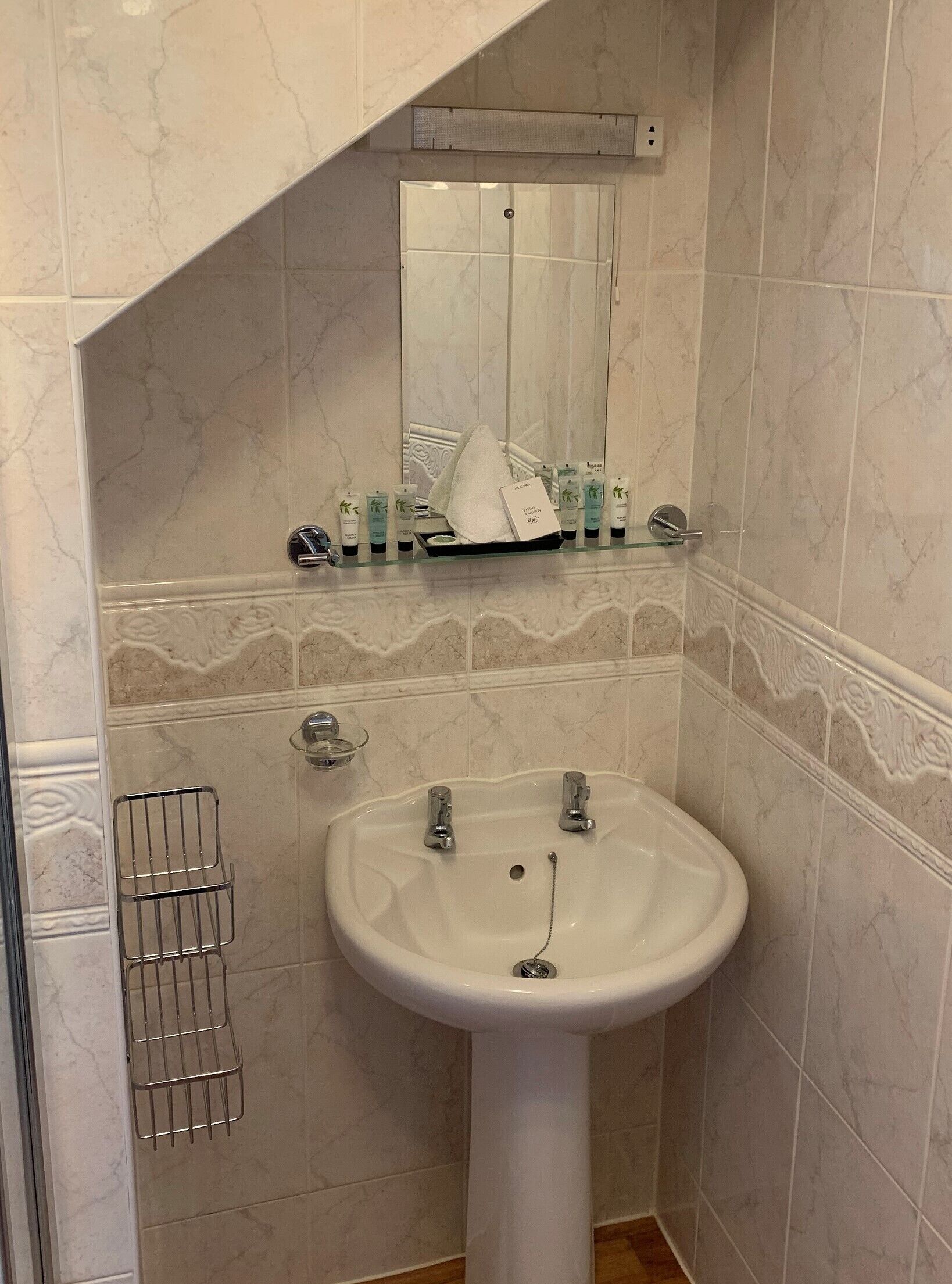 Un baño con lavamanos pedestal blanco, un espejo encima y un estante con artículos de aseo.