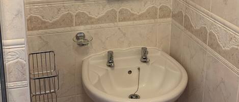 Un baño con lavamanos pedestal blanco, un espejo encima y un estante con artículos de aseo.