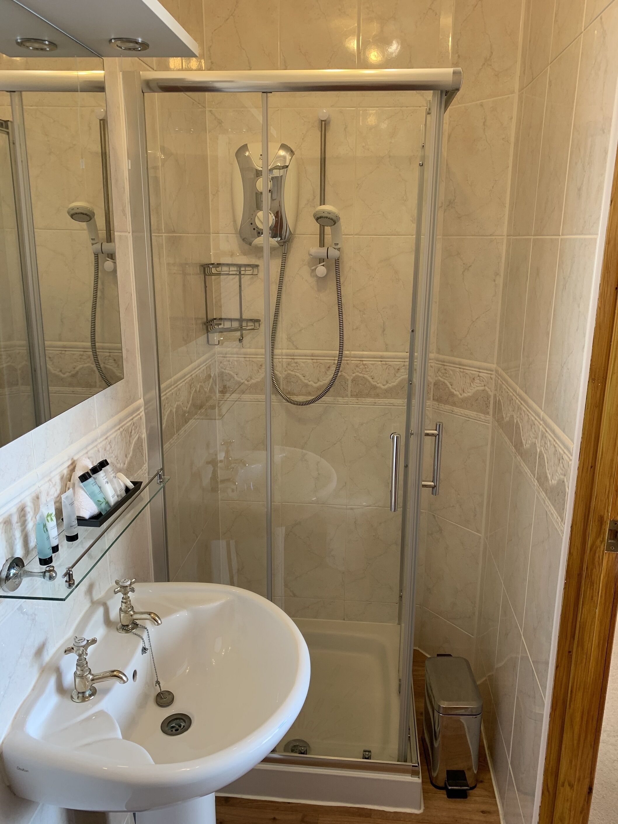 A glass-enclosed shower with a handheld showerhead and a wall-mounted faucet. A glass shelf with toiletries is mounted above the sink.