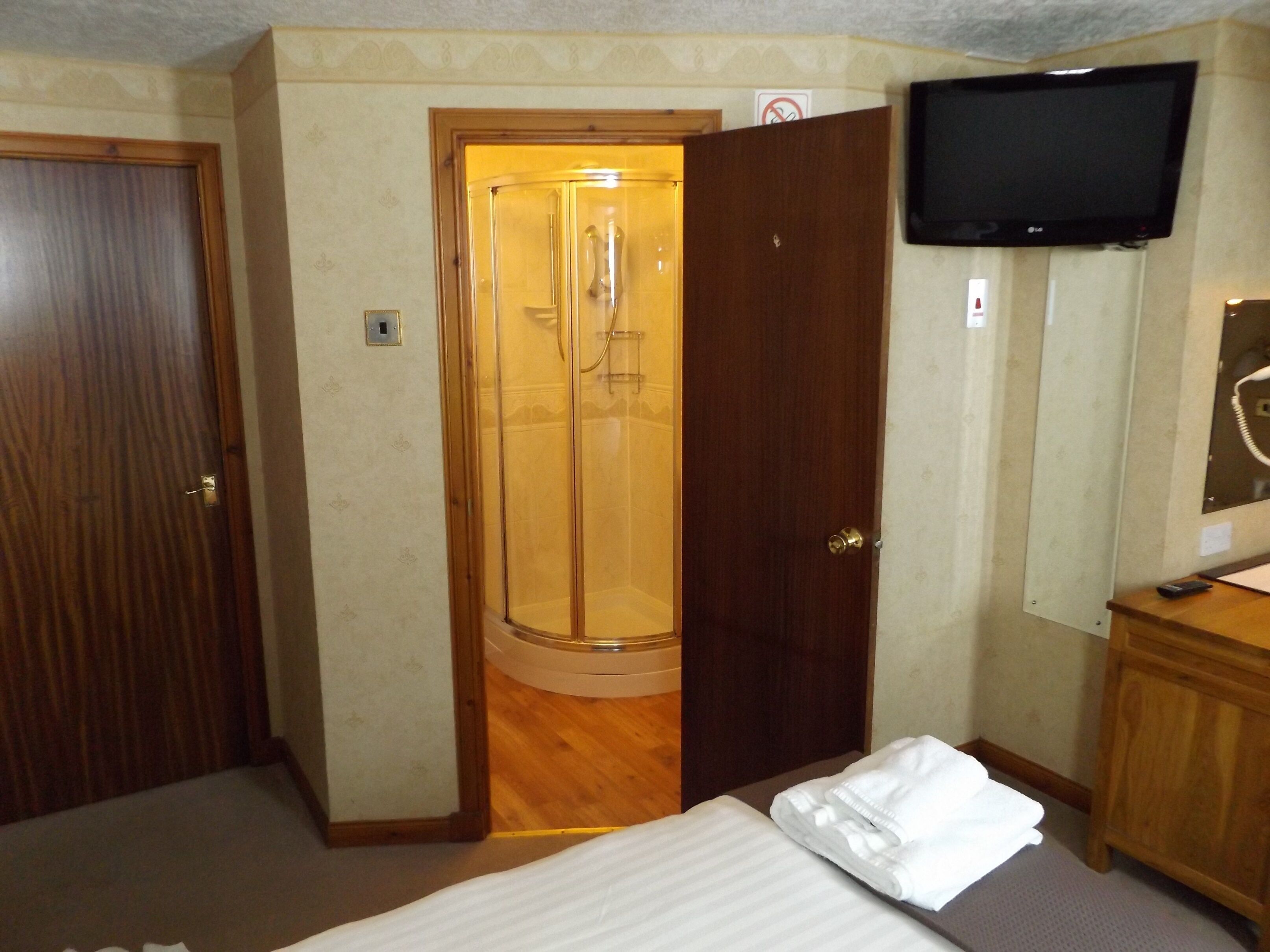 A hotel room with a wooden door, a glass shower enclosure, a bed with a white comforter, a wooden dresser, and a mounted television.