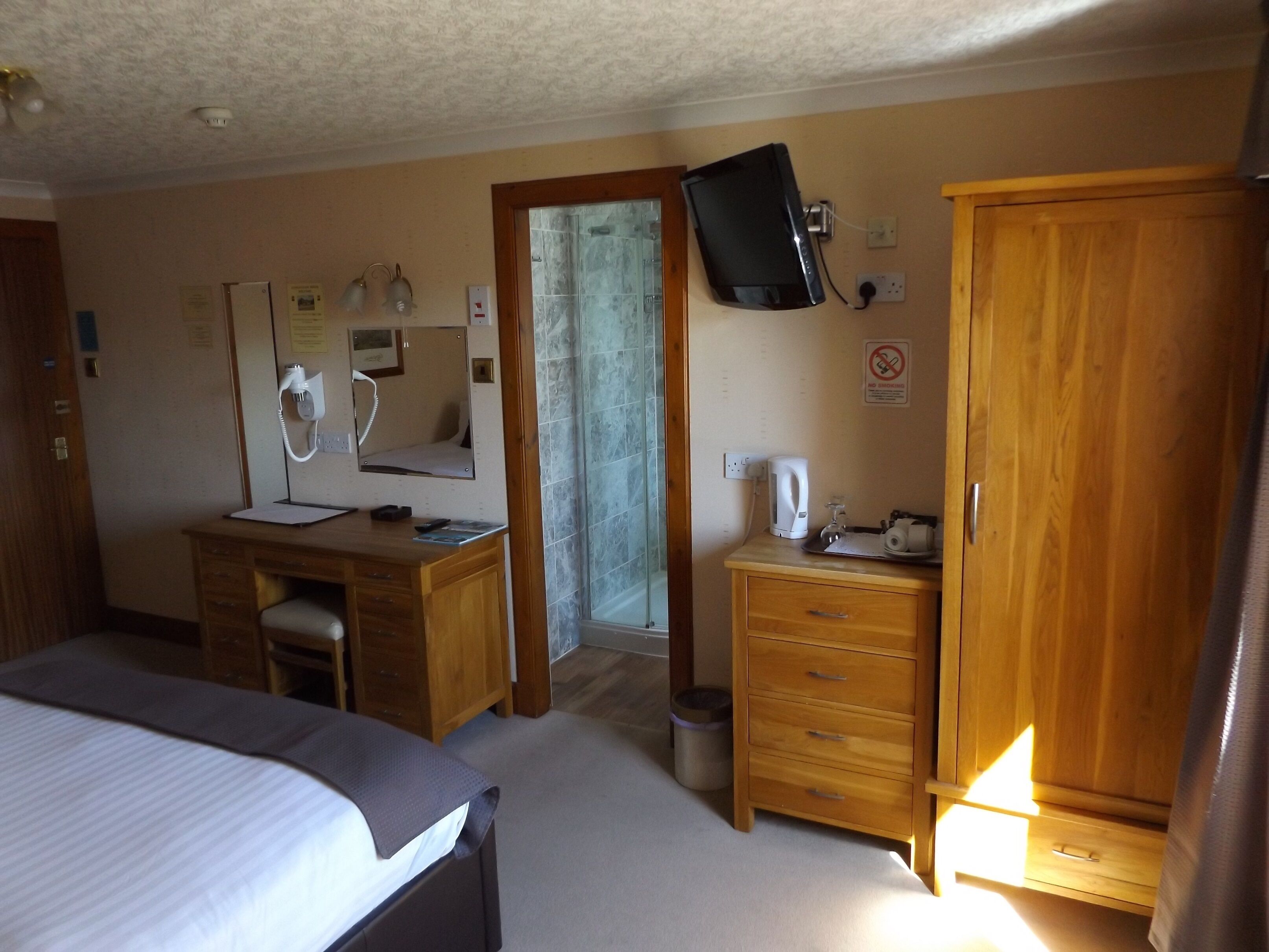 Habitación de hotel con cama, escritorio con teléfono, espejo, televisor en la pared, armario y pequeño mueble con tetera.