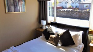 A hotel room with a bed, a nightstand, a lamp, a painting, and a window with a view of houses. - Lyndon Guest House (Inverness)