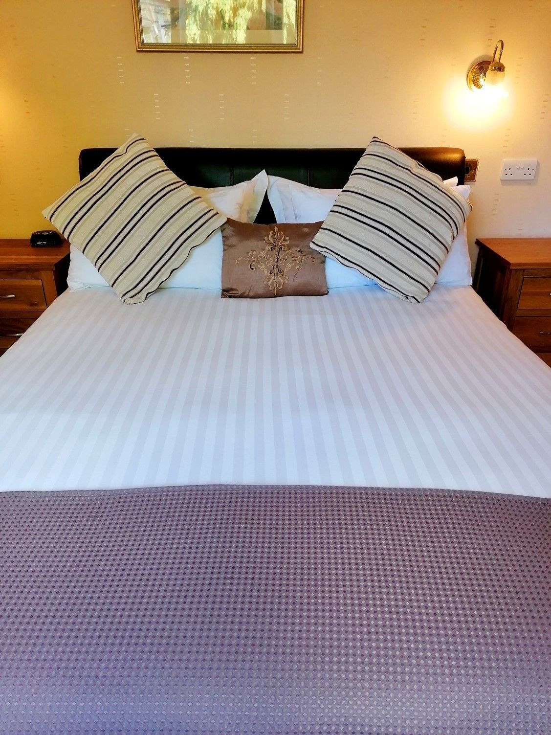 A neatly made bed with striped pillows and a decorative central pillow, a wooden nightstand with a lamp, and a wall-mounted light fixture.