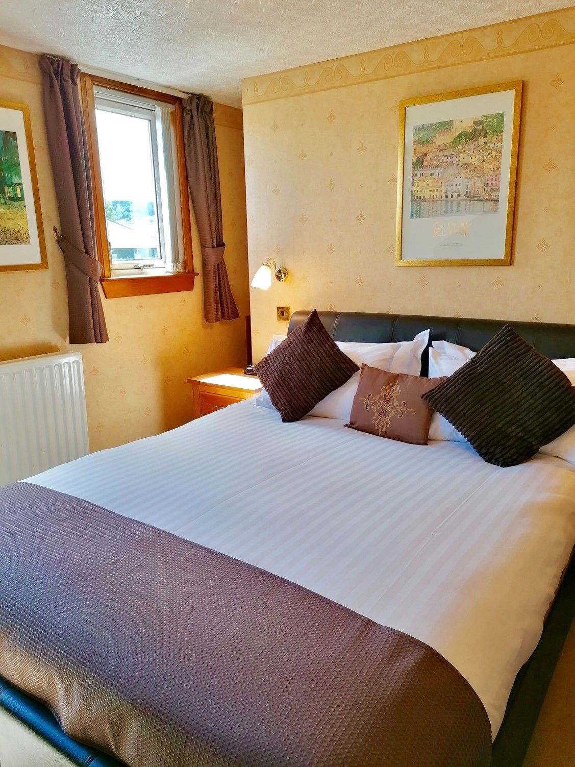A hotel room with a large bed, a nightstand with a lamp, a window with curtains, and two framed pictures on the wall.