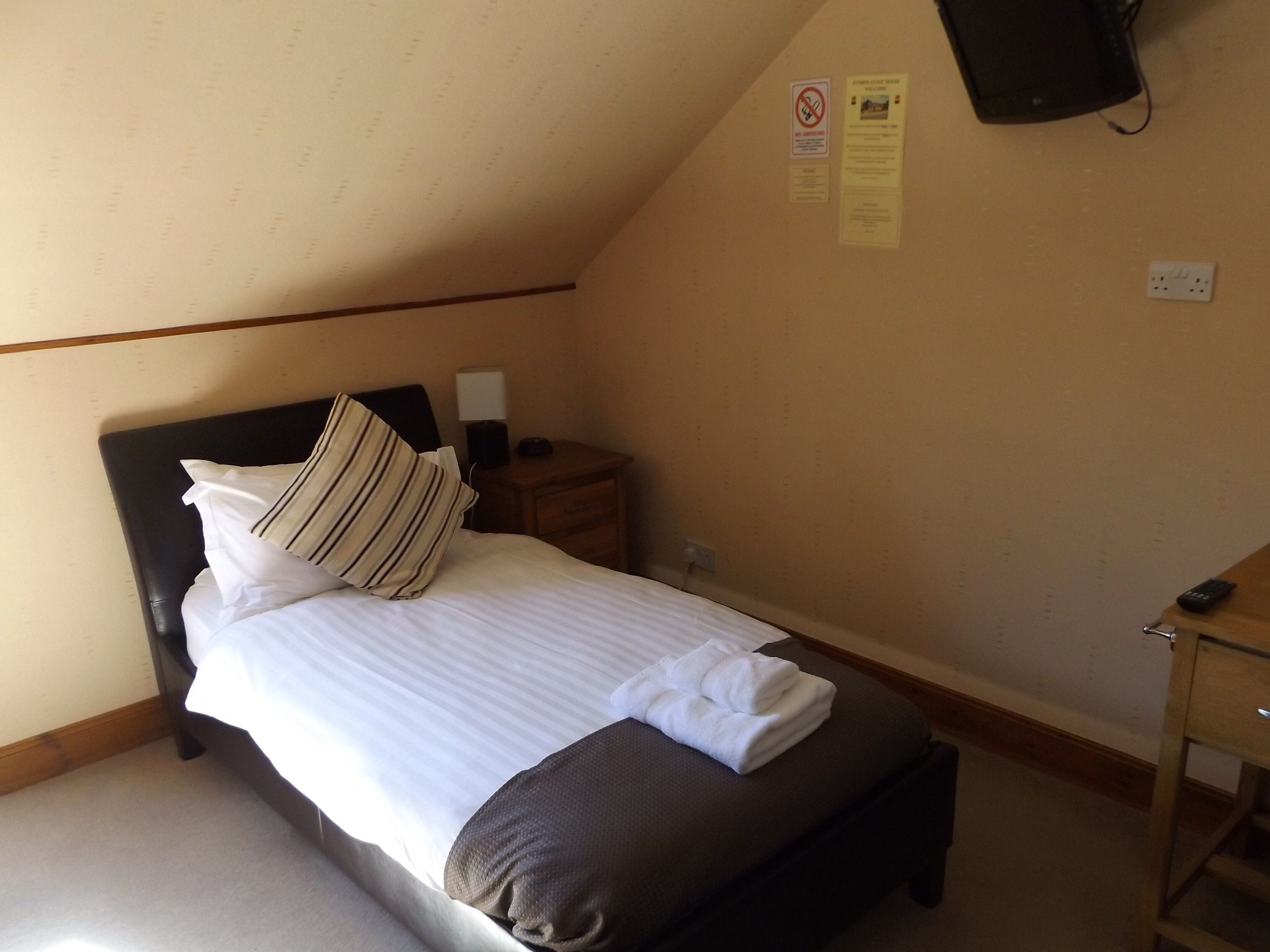 A neatly made bed with pillows and a folded towel, a bedside table with a lamp, a wall-mounted TV, and a power outlet.