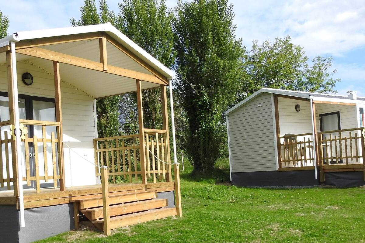 Two mobile homes with wooden decks and porches, surrounded by green grass and trees.