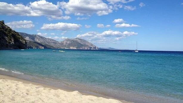 Una playa con aguas turquesas cristalinas, orilla de arena y montañas al fondo.
