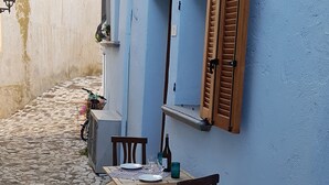 Un estrecho callejón adoquinado con una mesa dispuesta para dos, sillas de madera y una pared azul con una ventana y persiana.