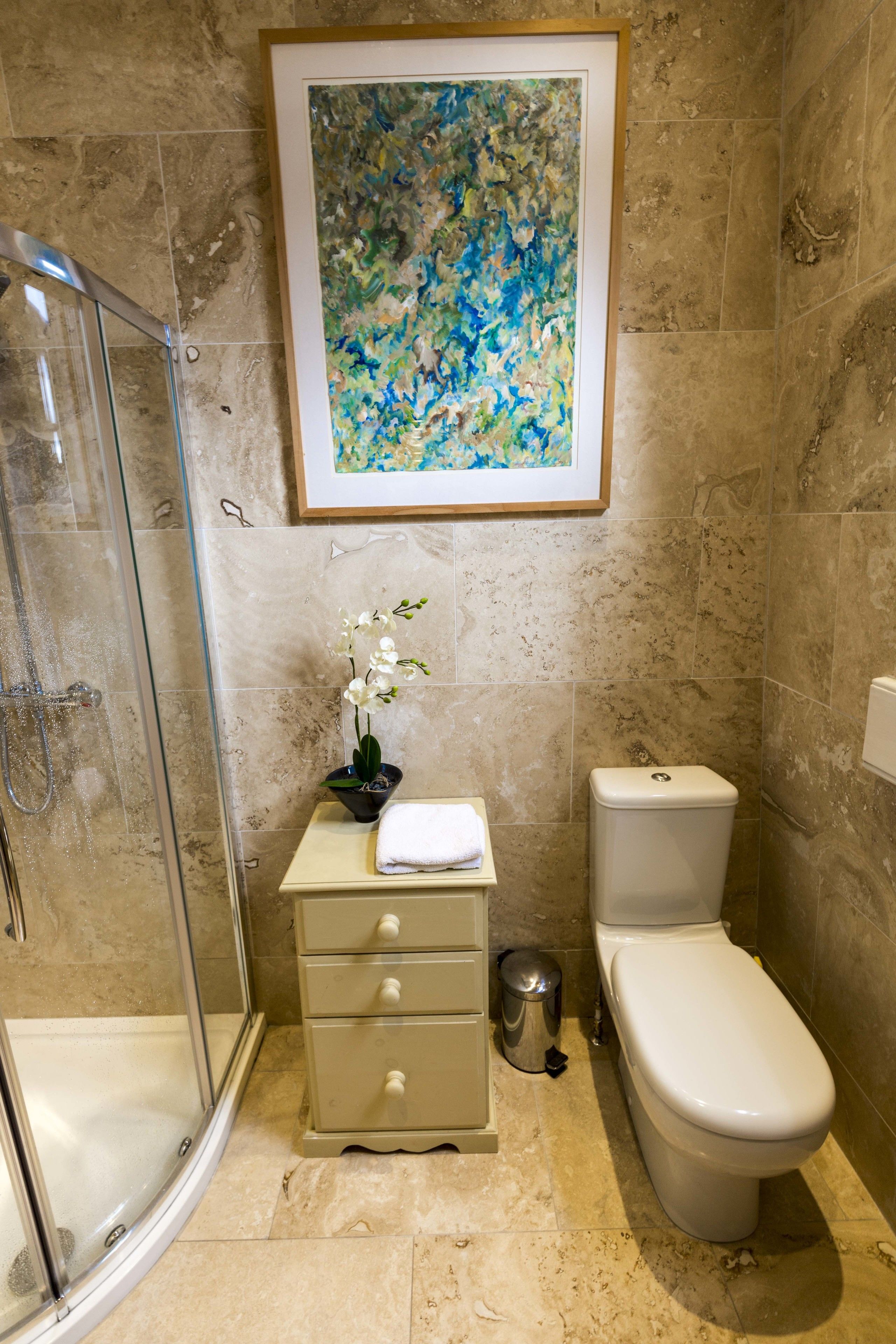 A bathroom with a glass shower enclosure, a toilet, a sink with a towel, and a framed abstract painting on the wall.