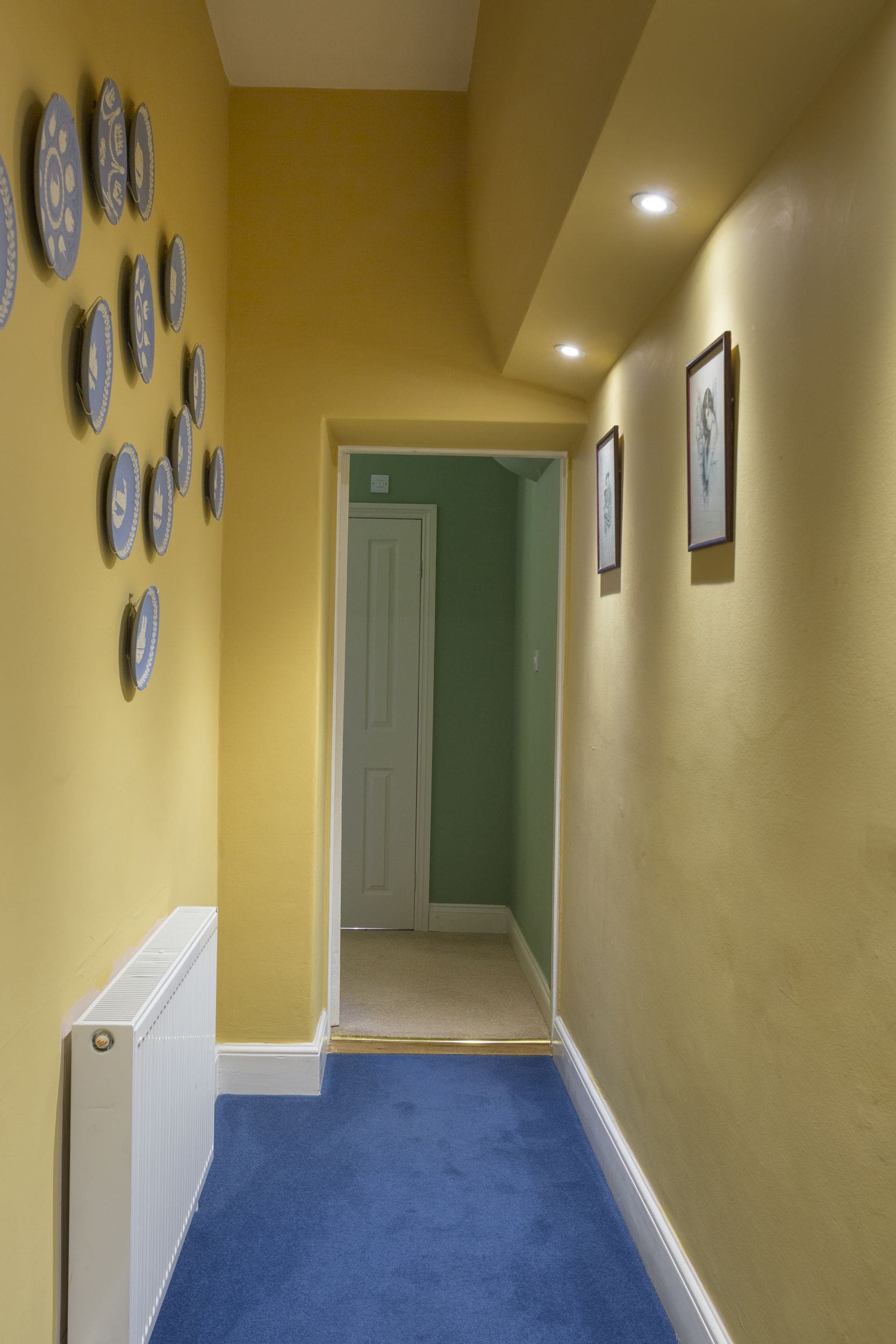 Un couloir avec un tapis bleu, des murs jaunes et un radiateur blanc.