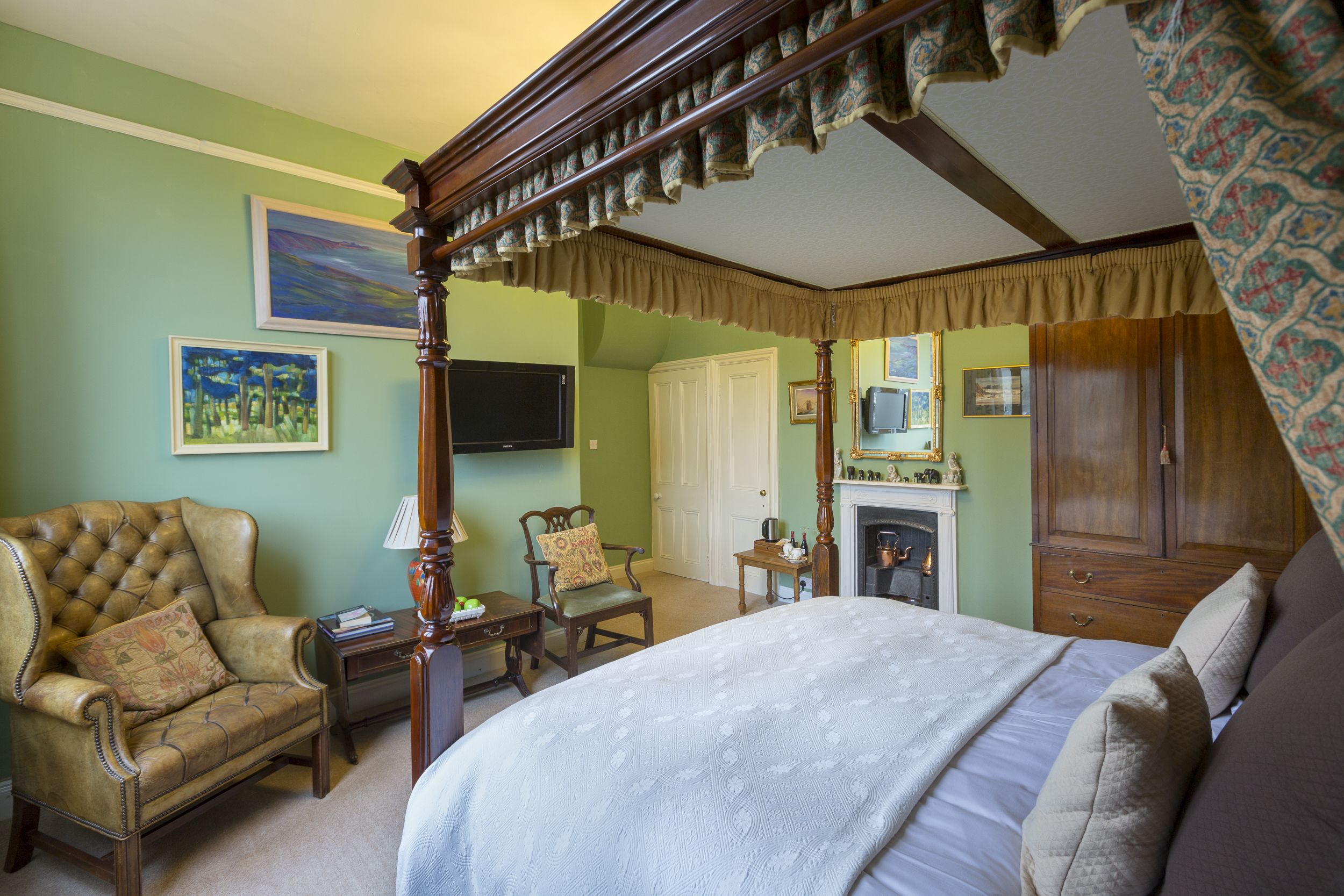 A bedroom with a four-poster bed, a sitting area with a chair and a small table, a fireplace, and a wardrobe.