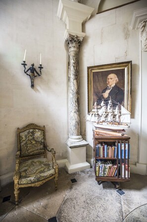 Una habitación con una columna de mármol, un retrato enmarco, una silla decorativa y una estantería.