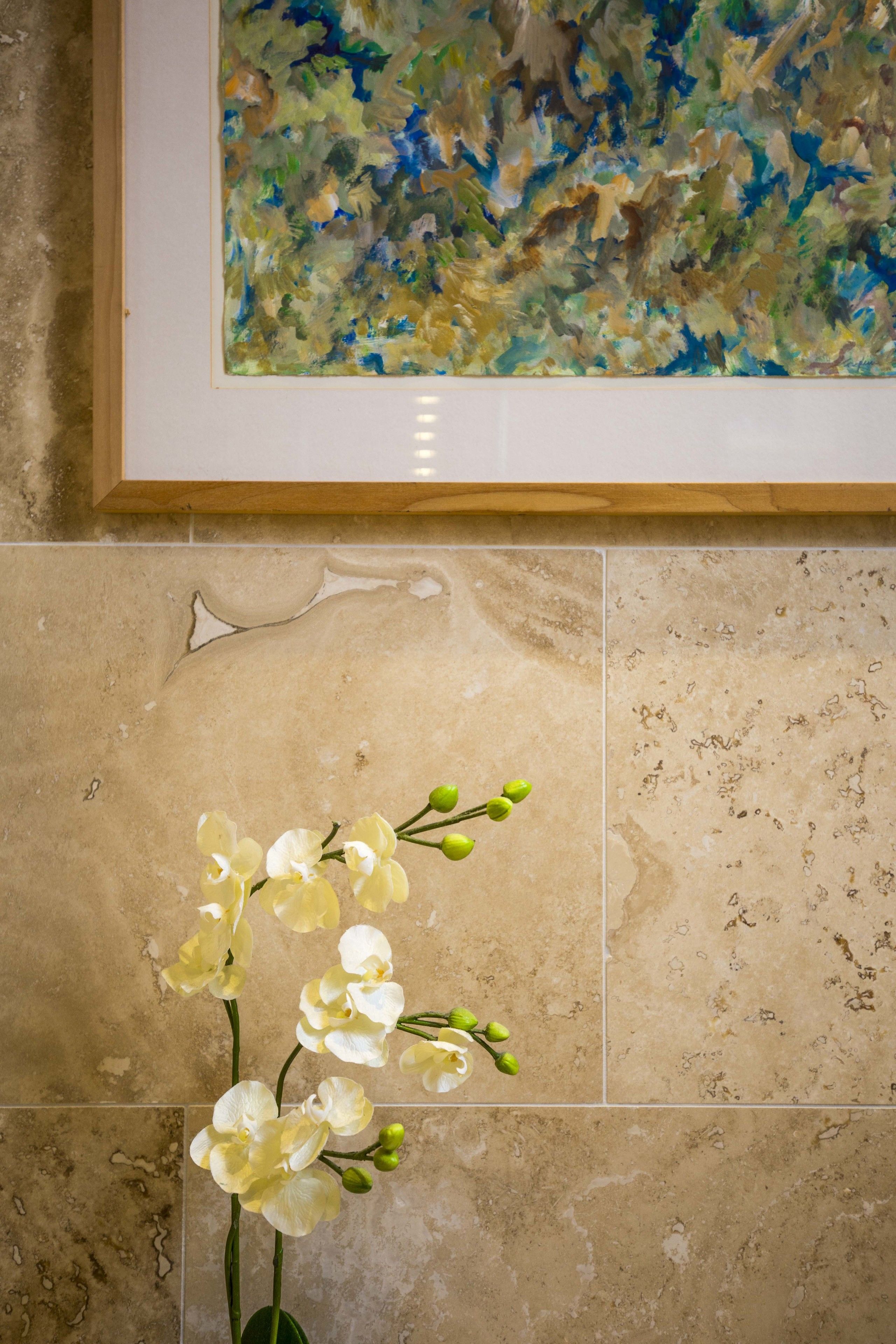 A framed abstract painting above a tiled wall with a vase of white orchids.