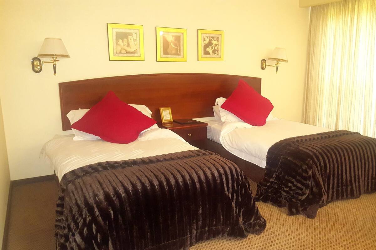 Habitación de hotel con dos camas, cada una con almohada roja y colcha negra, cabecero de madera y mesita de noche con lámpara. Se observan tres cuadros enmarcados en la pared sobre las camas.