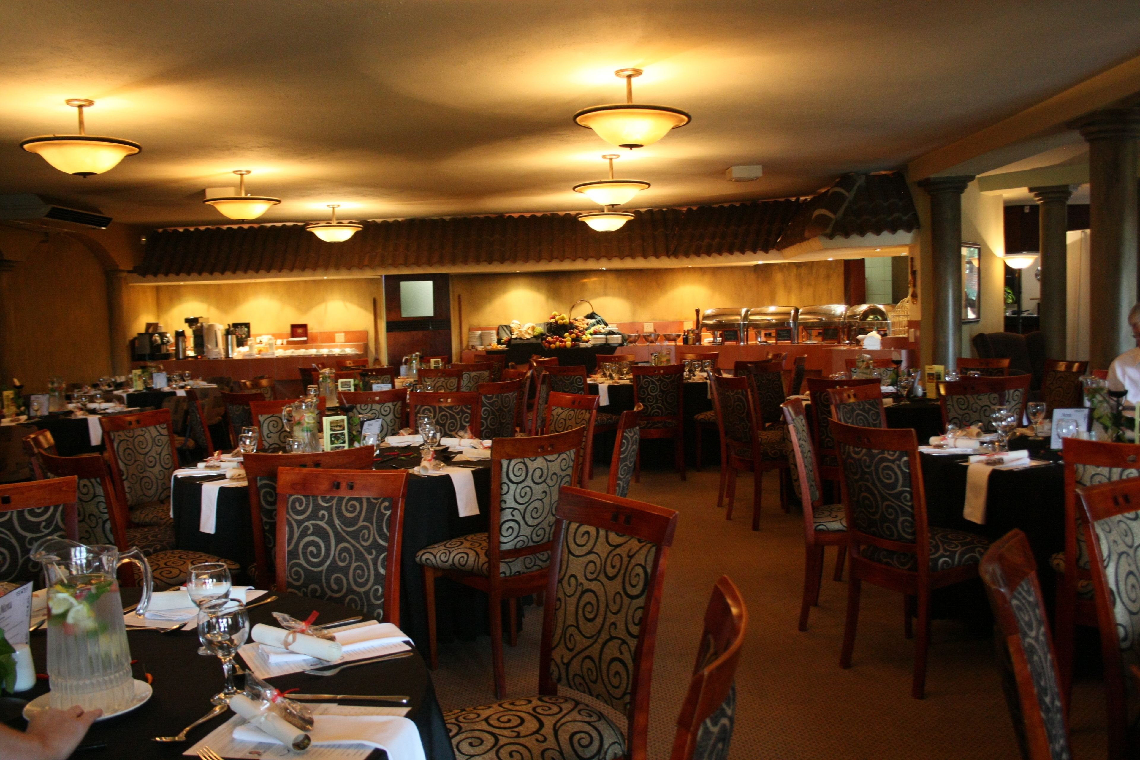 En restaurant med flere spiseborde og stole, pendellamper og et buffetbord i baggrunden.