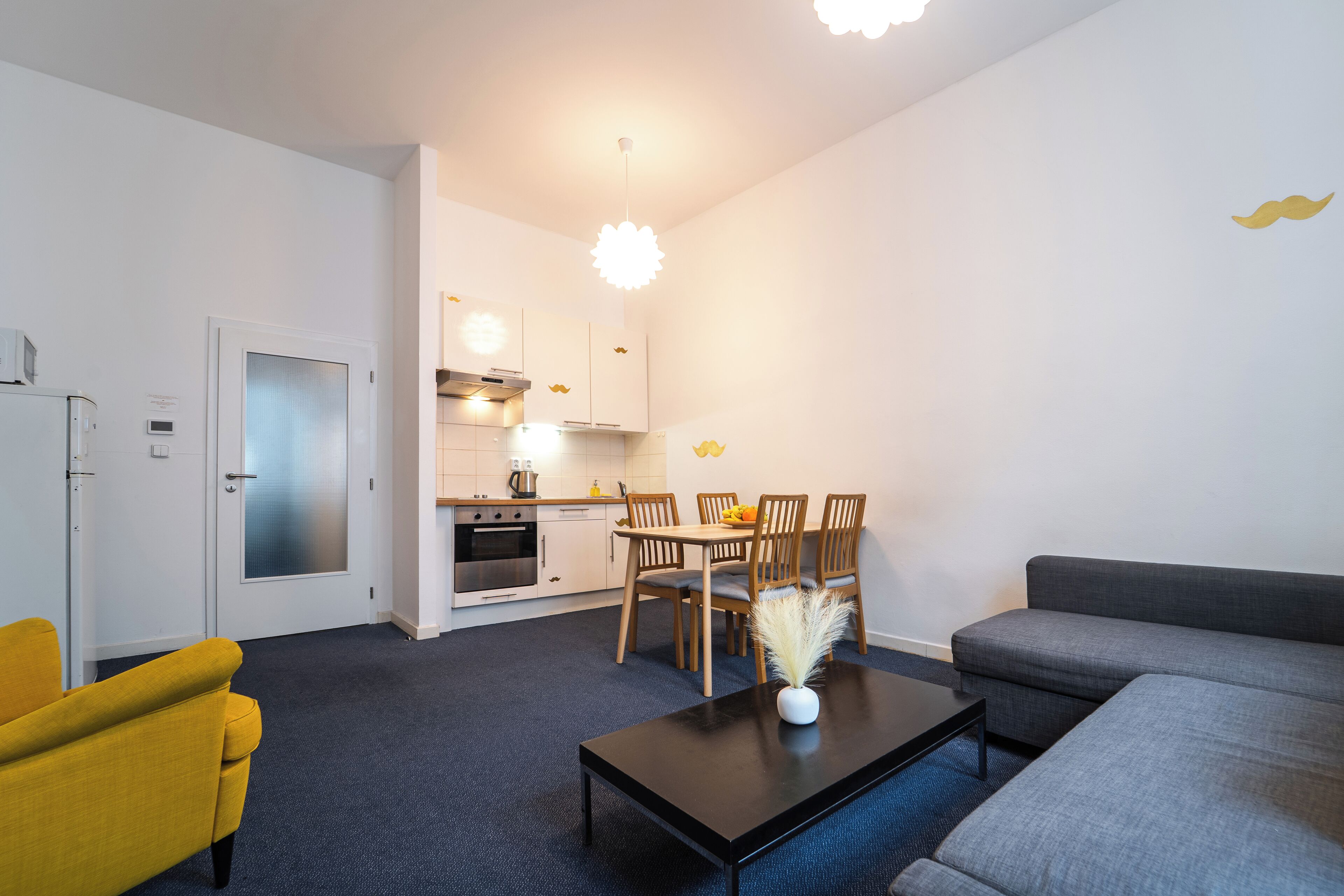A modern living room with a sofa, a yellow armchair, a dining table with chairs, and a kitchen in the background.