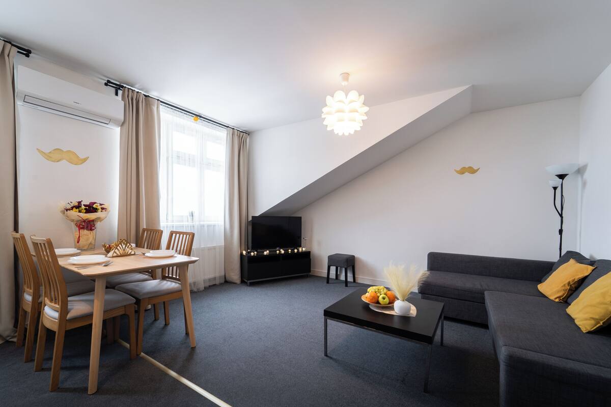 Uma sala de estar moderna com área de jantar, um sofá preto, uma televisão e uma luminária de teto diferenciada.