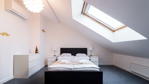 Un dormitorio moderno con tragaluces, una cama con ropa de cama blanca, una mesita de noche con lámpara y un aparador.